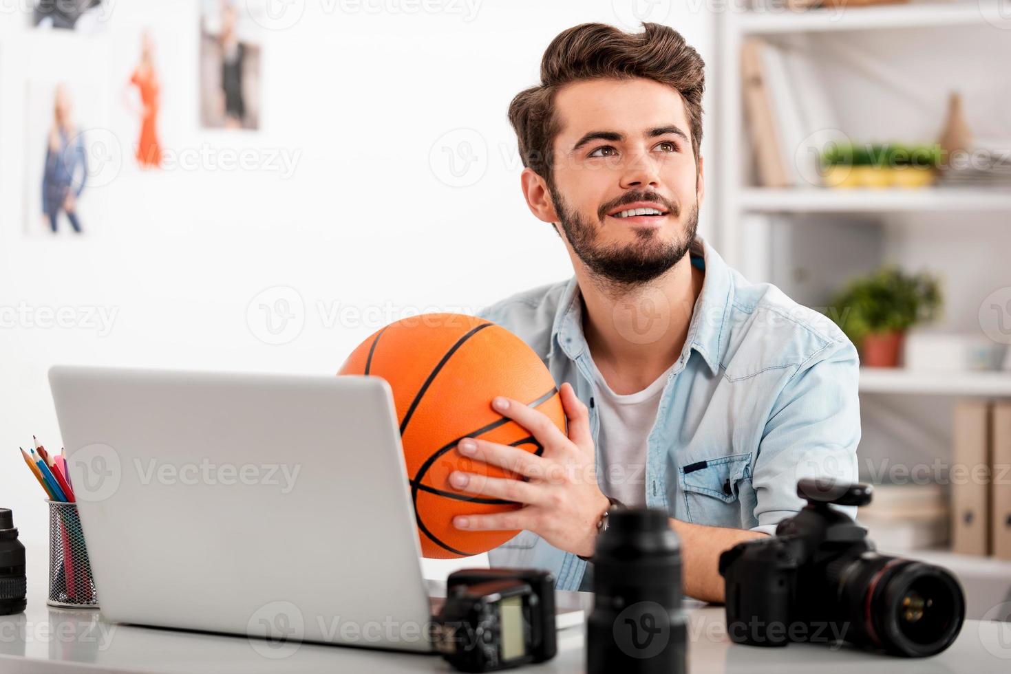 immaginazione è un' chiave per successo. contento giovane uomo Tenere pallacanestro palla e guardare lontano mentre seduta a il suo Lavorando posto foto