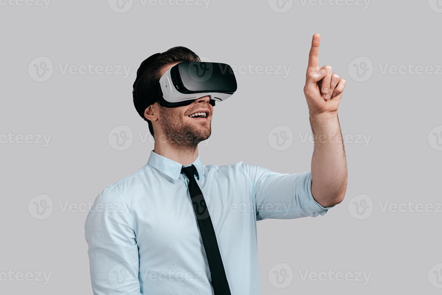 godendo innovativo tecnologie. bene guardare giovane uomo nel virtuale la realtà cuffia puntamento nel il aria mentre in piedi contro grigio sfondo foto