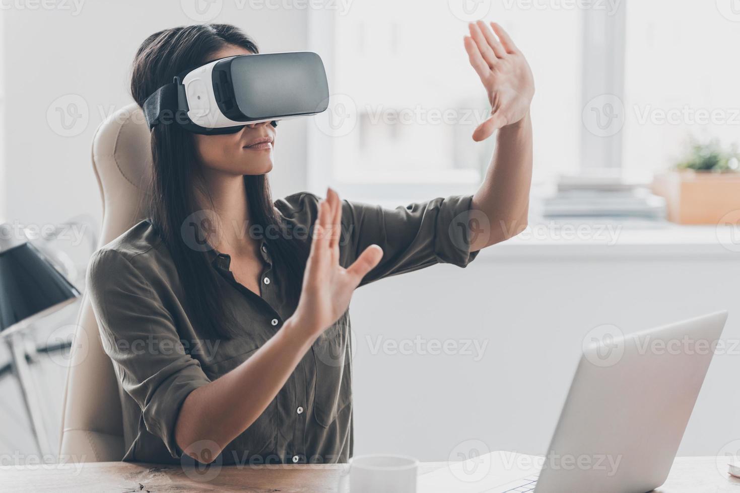 donna con occhiali vr. giovane donna sicura di sé in cuffia per realtà virtuale che punta in aria mentre è seduta al suo posto di lavoro in ufficio foto