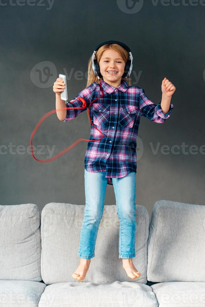 gioia e felicità. bellissimo poco nel cuffie Tenere sua inteligente Telefono e guardare a telecamera con Sorridi mentre salto su il divano a casa foto