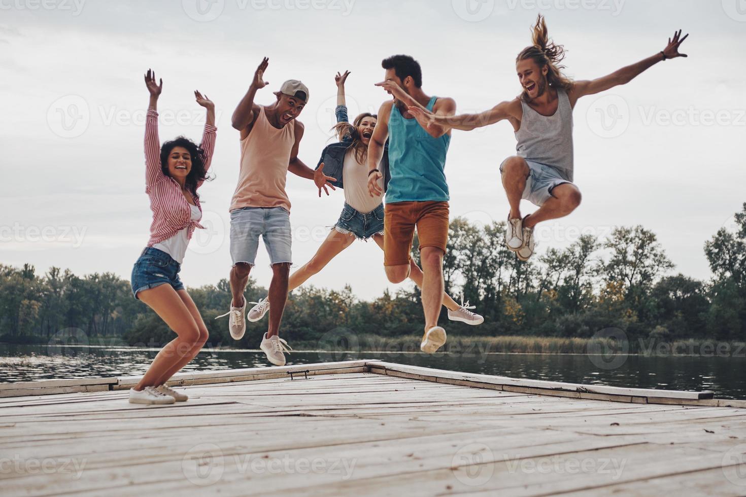 grande giorno con gli amici. pieno lunghezza di giovane persone nel casuale indossare sorridente e Esprimere a gesti mentre salto su il molo foto