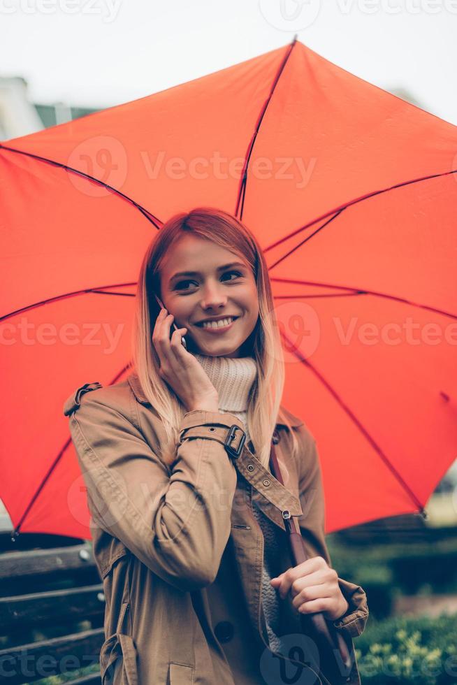 simpatico parlare con amico. attraente giovane sorridente donna trasporto ombrello e parlando su il mobile Telefono mentre seduta su il panchina all'aperto foto