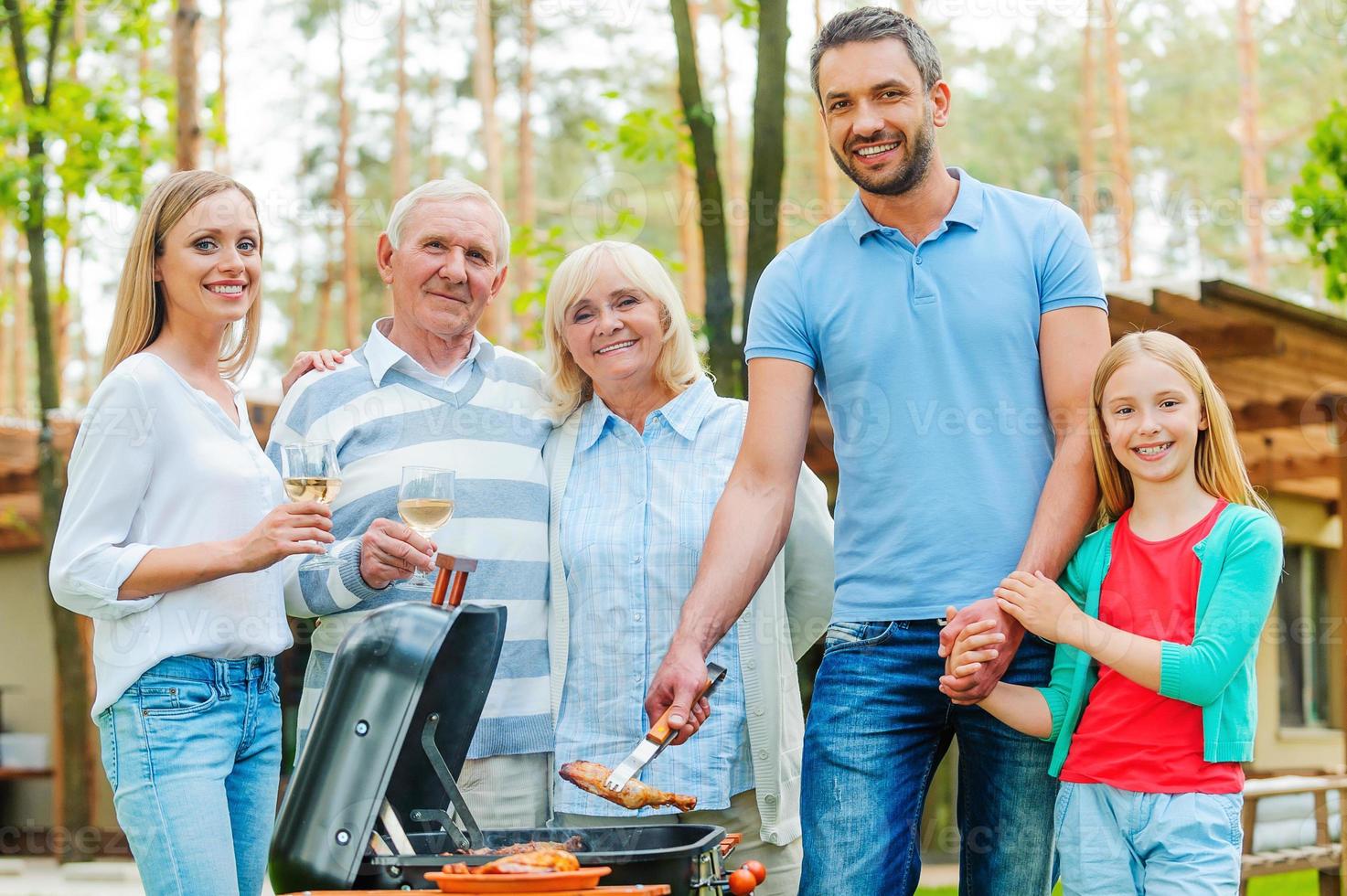 barbeque volta. contento famiglia di cinque persone barbecue carne su griglia e guardare a telecamera foto