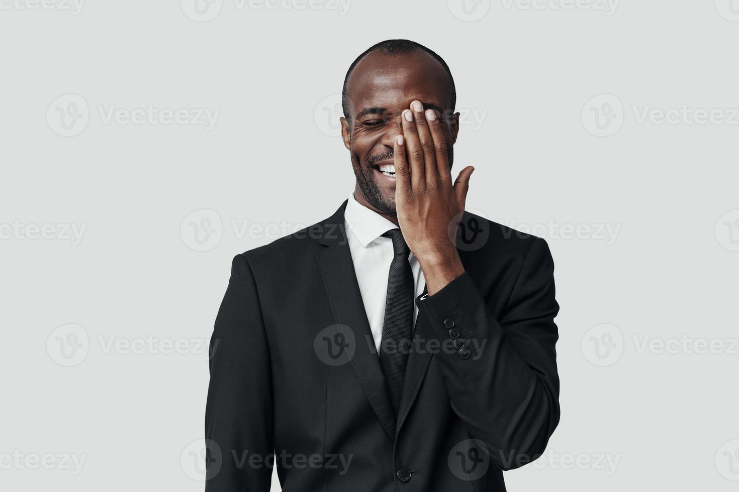 giocoso giovane africano uomo nel vestito formale copertura metà di il suo viso con mano e sorridente mentre in piedi contro grigio sfondo foto