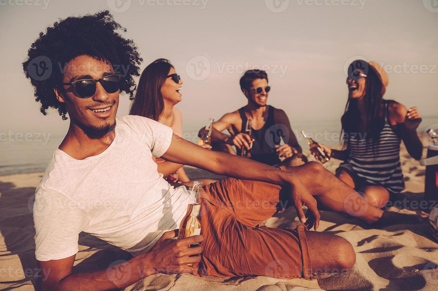 godendo spiaggia festa con gli amici. allegro giovane persone la spesa simpatico tempo insieme mentre seduta su il spiaggia e potabile birra foto