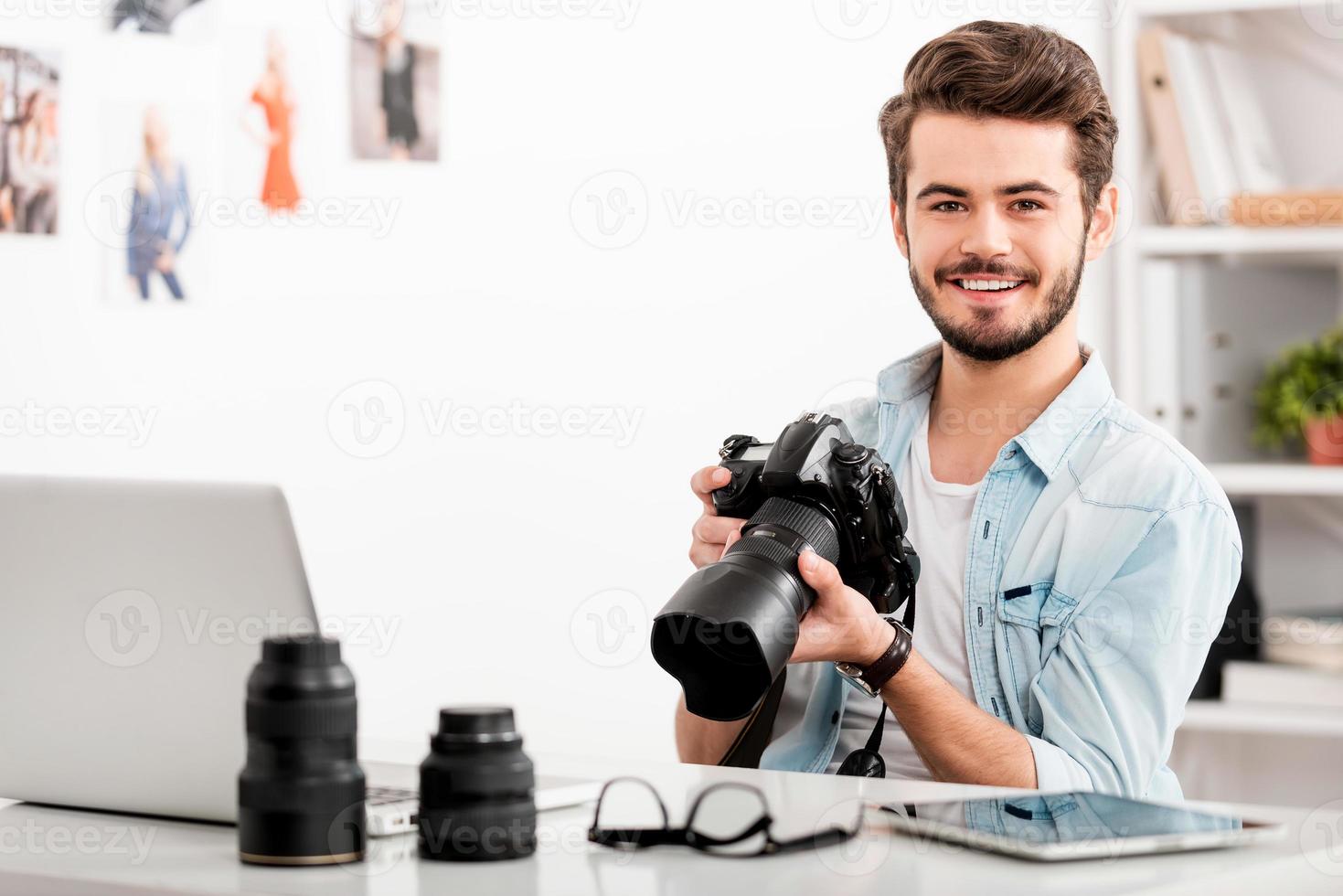 fiducioso fotografo. allegro giovane uomo Tenere digitale telecamera e sorridente mentre seduta a il suo Lavorando posto foto