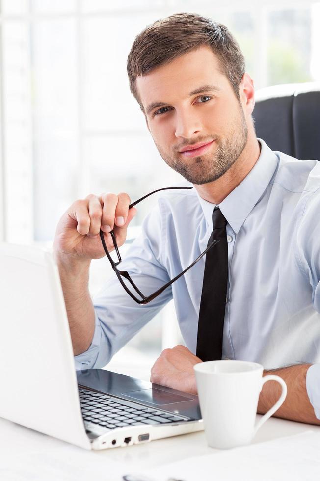 fabbricazione il tuo attività commerciale Più facile. fiducioso giovane uomo nel vestito formale Tenere bicchieri e guardare a telecamera mentre seduta a il suo Lavorando posto foto