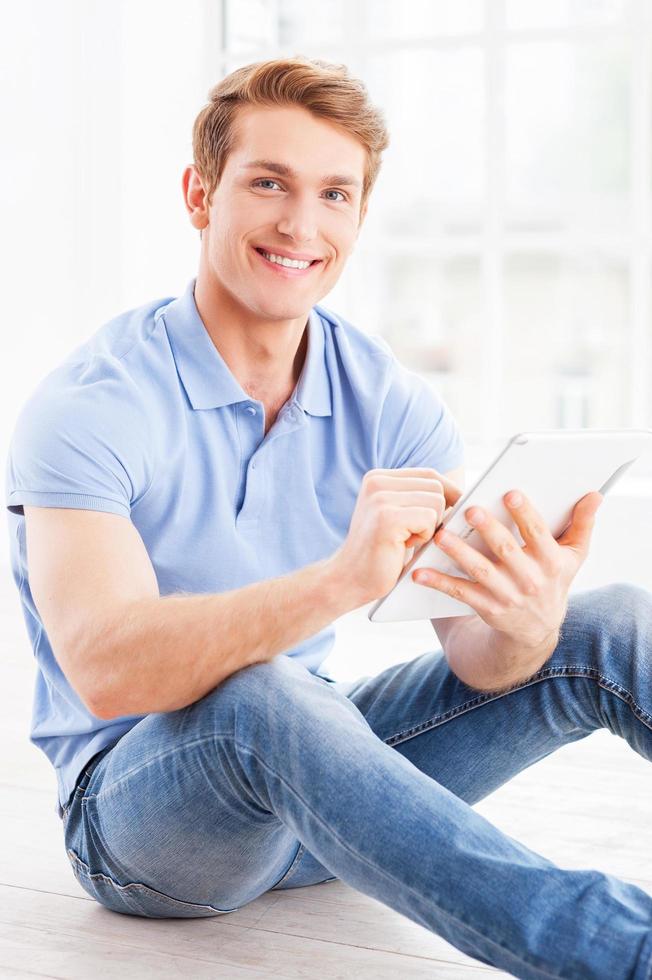 l'esame il suo marca nuovo tavoletta. bello giovane uomo Lavorando su digitale tavoletta e sorridente mentre seduta su il pavimento a il suo appartamento foto
