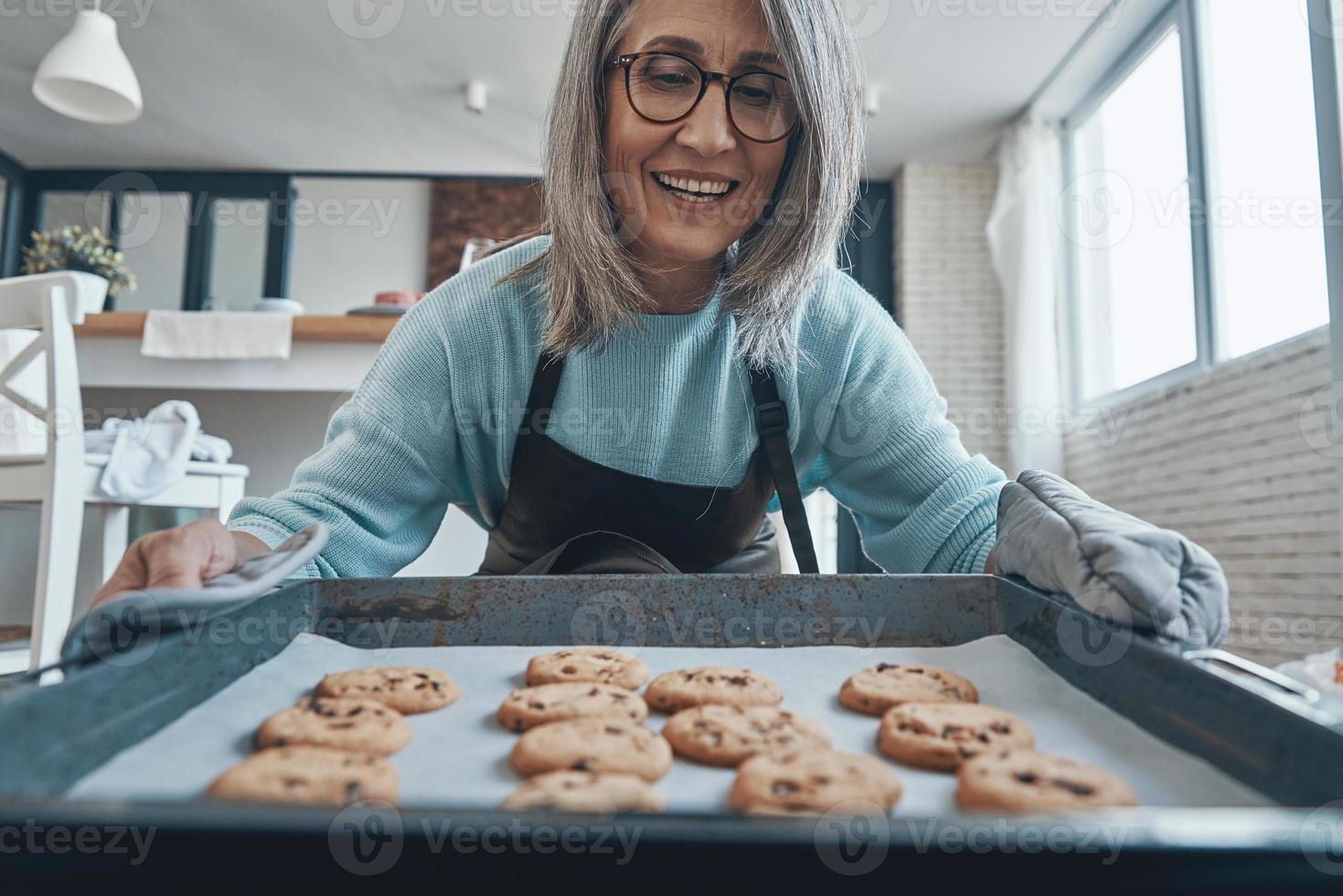 contento anziano donna sorridente mentre preparazione biscotti nel il cucina foto