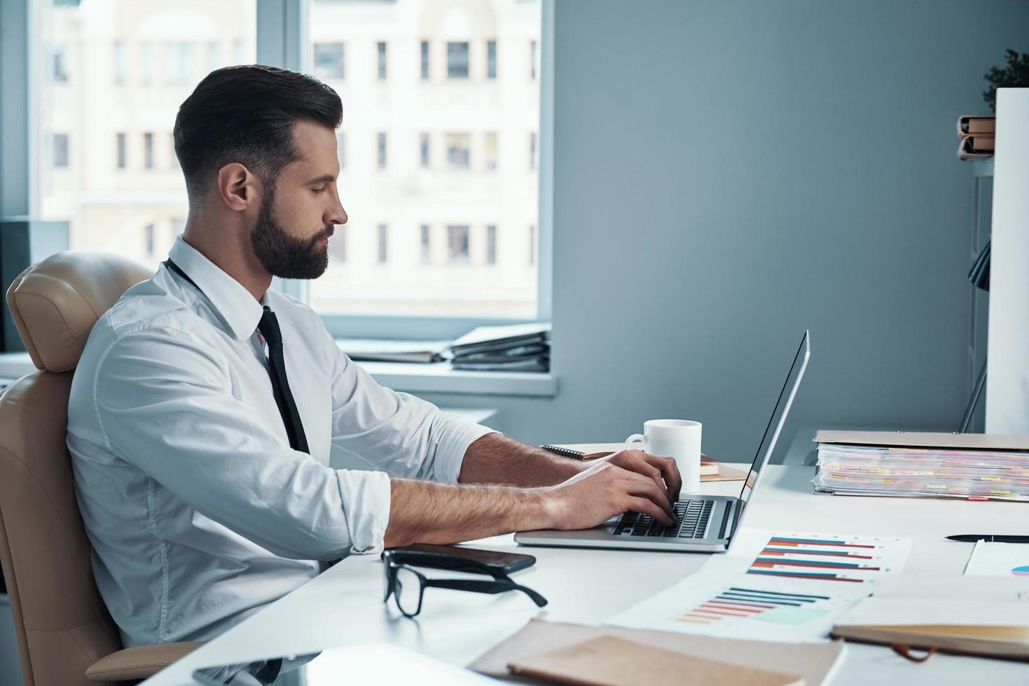 concentrato giovane uomo nel camicia e cravatta Lavorando su computer mentre seduta nel il ufficio foto