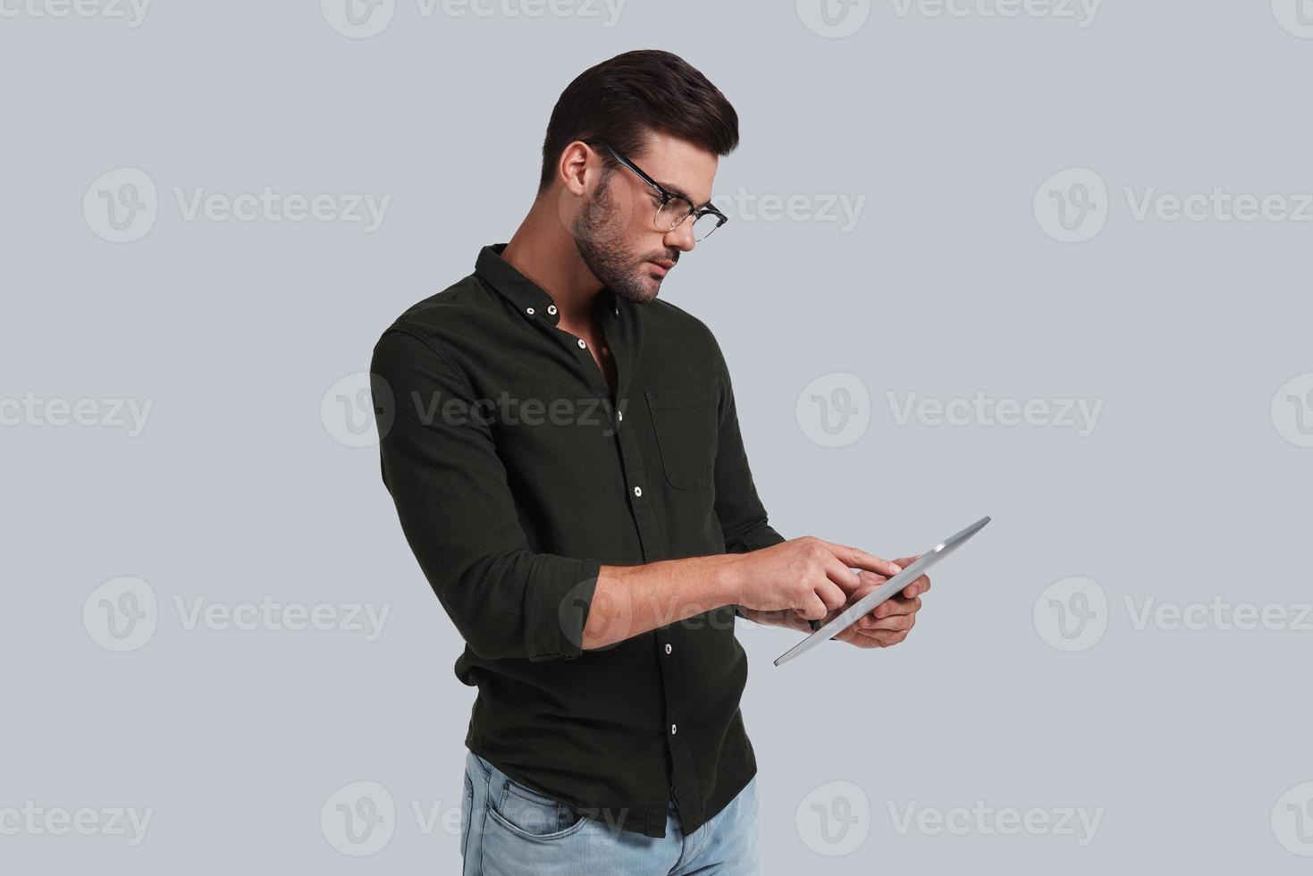 l'esame il suo digitale tavoletta. grave giovane uomo nel occhiali Lavorando su il suo digitale tavoletta mentre in piedi contro grigio sfondo foto
