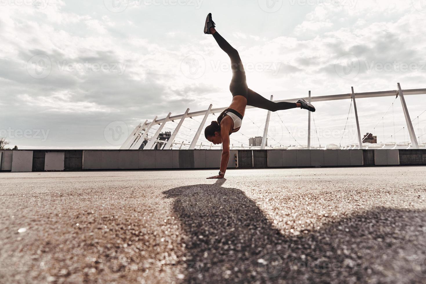 energia e equilibrio. pieno lunghezza di moderno giovane donna nel gli sport capi di abbigliamento fare handstand mentre esercizio all'aperto foto