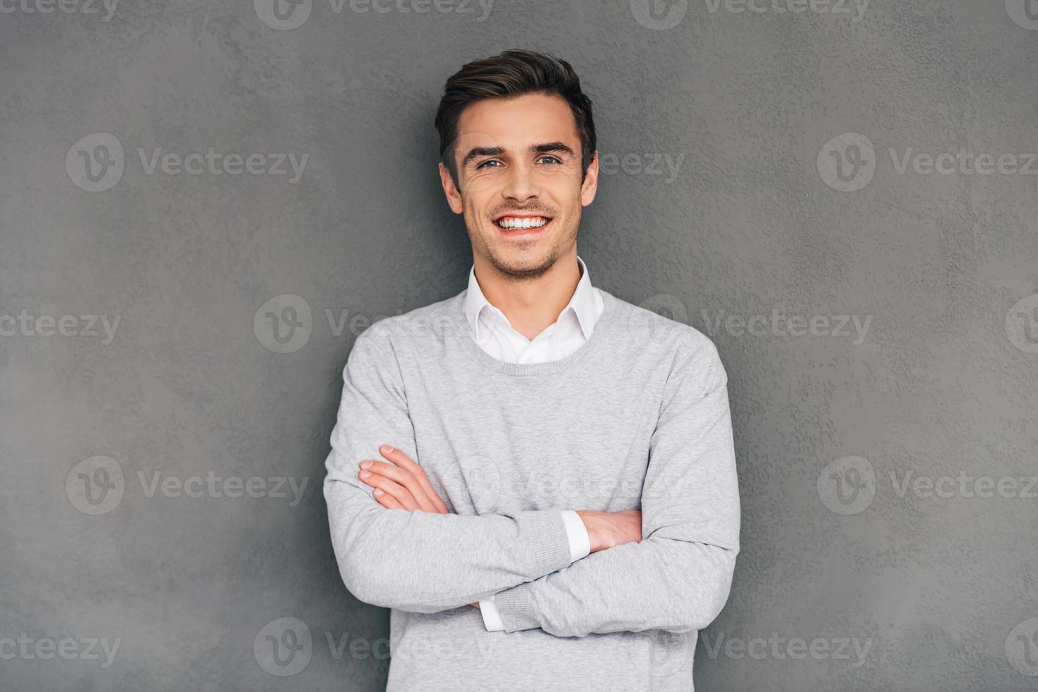 mantenere sorridente. fiducioso giovane uomo conservazione braccia attraversato e guardare a telecamera con Sorridi mentre in piedi contro grigio sfondo foto