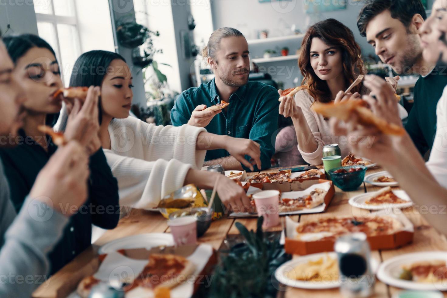 condivisione grande pasto. gruppo di giovane persone nel casuale indossare mangiare Pizza e sorridente mentre avendo un' cena festa in casa foto