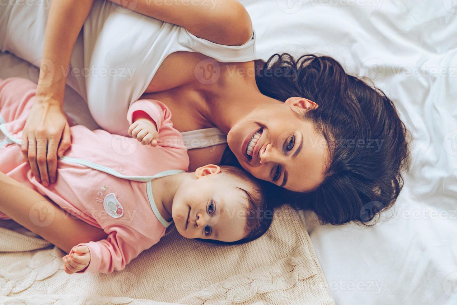 mattina abbracci. superiore Visualizza di allegro bellissimo giovane donna guardare a telecamera con Sorridi mentre dire bugie nel letto con sua bambino ragazza foto