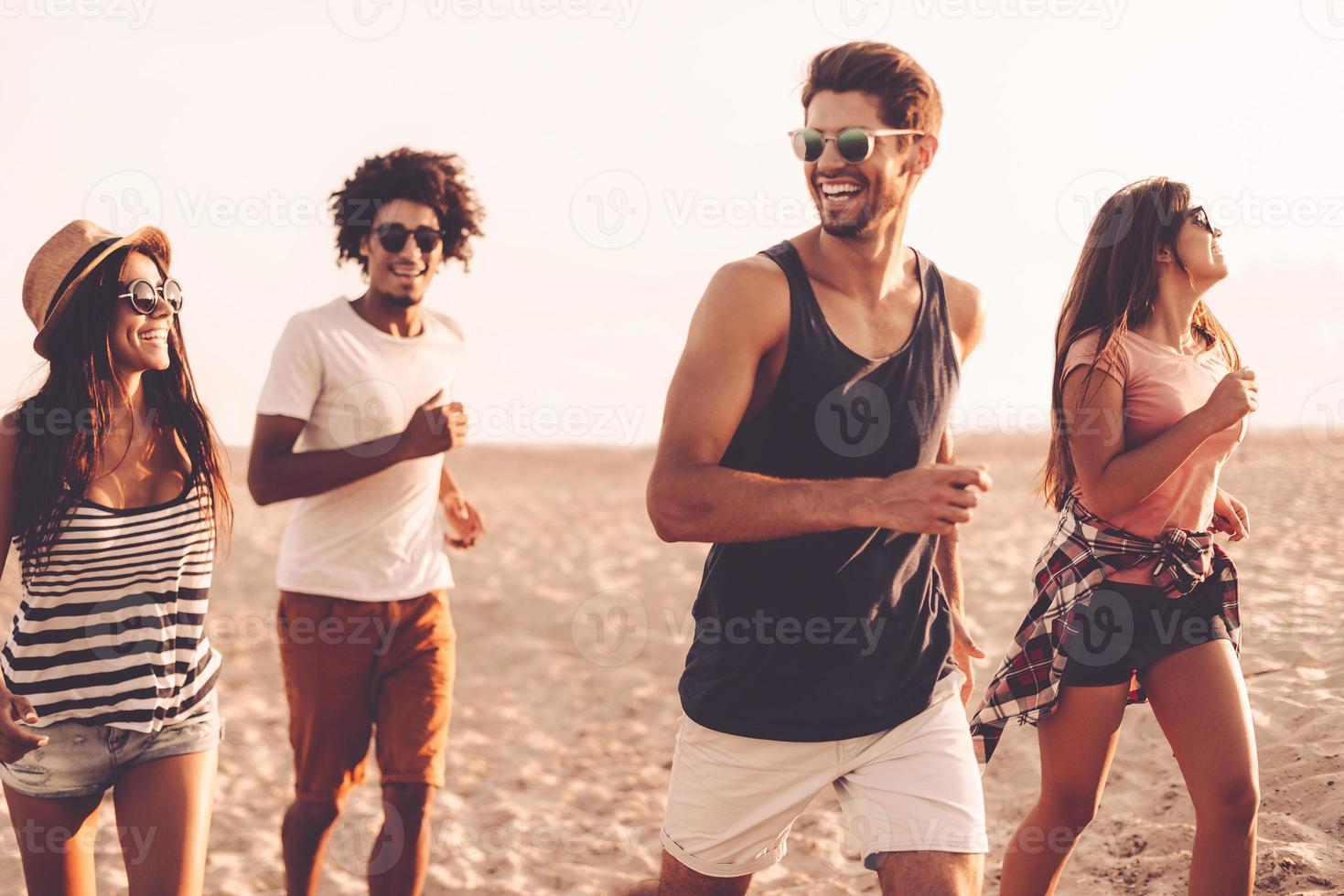 godendo libertà. gruppo di giovane allegro persone in esecuzione lungo il spiaggia e guardare contento foto