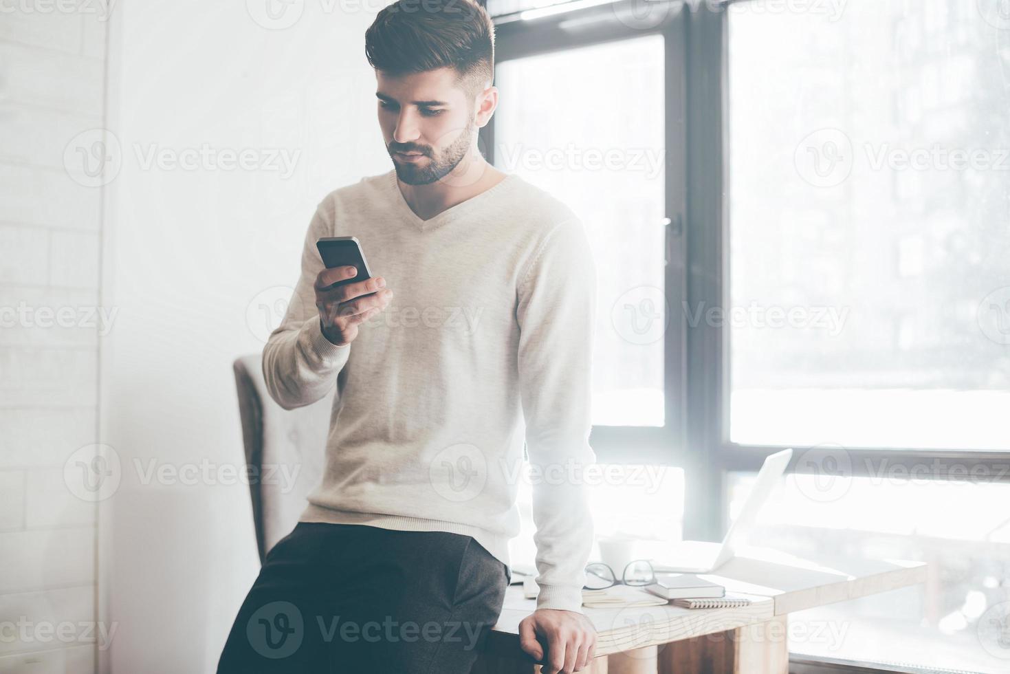 digitando Presto Messaggio. fiducioso giovane uomo Tenere inteligente Telefono e guardare a esso mentre in piedi vicino il suo Lavorando posto nel ufficio foto