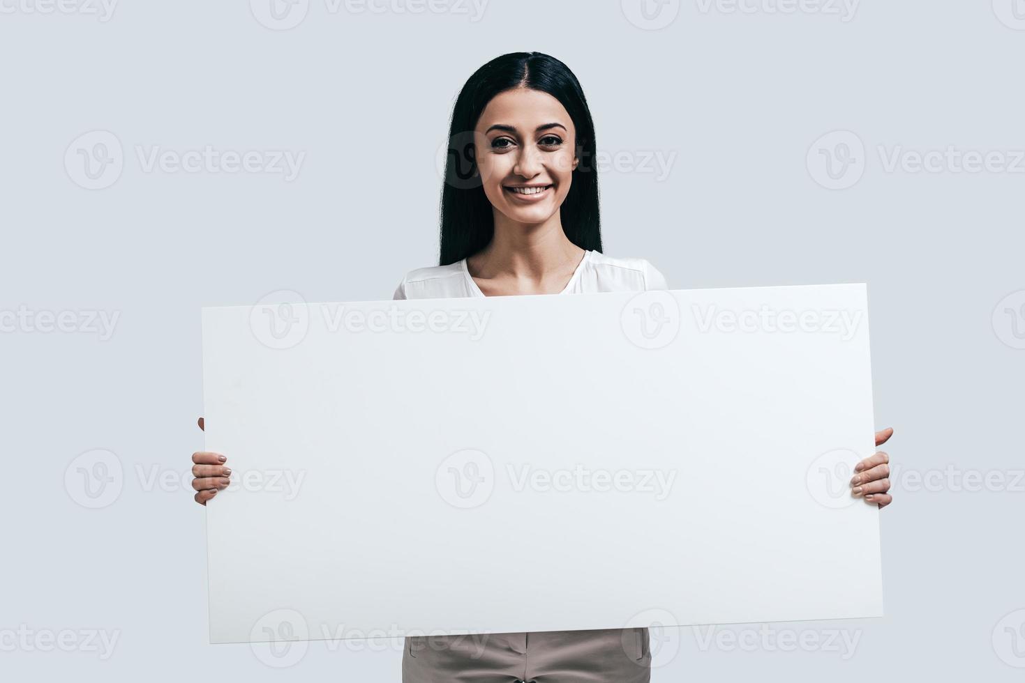 bellissimo e pieno di energia. giovane fiducioso donna Tenere vuoto flip chart e guardare a telecamera mentre in piedi contro grigio sfondo foto