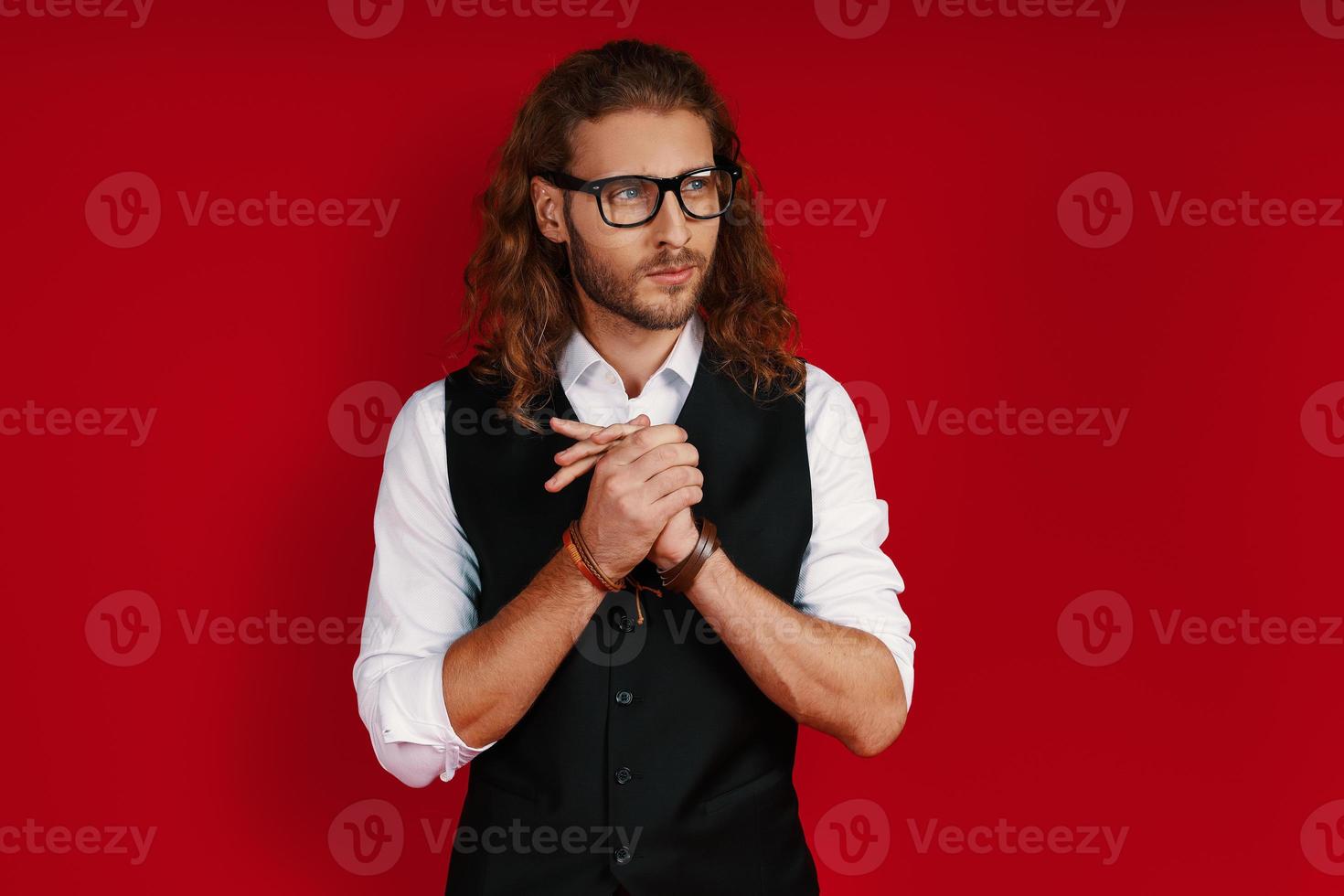 riflessivo giovane uomo nel elegante capi di abbigliamento guardare lontano e conservazione mani stretto mentre in piedi contro rosso sfondo foto