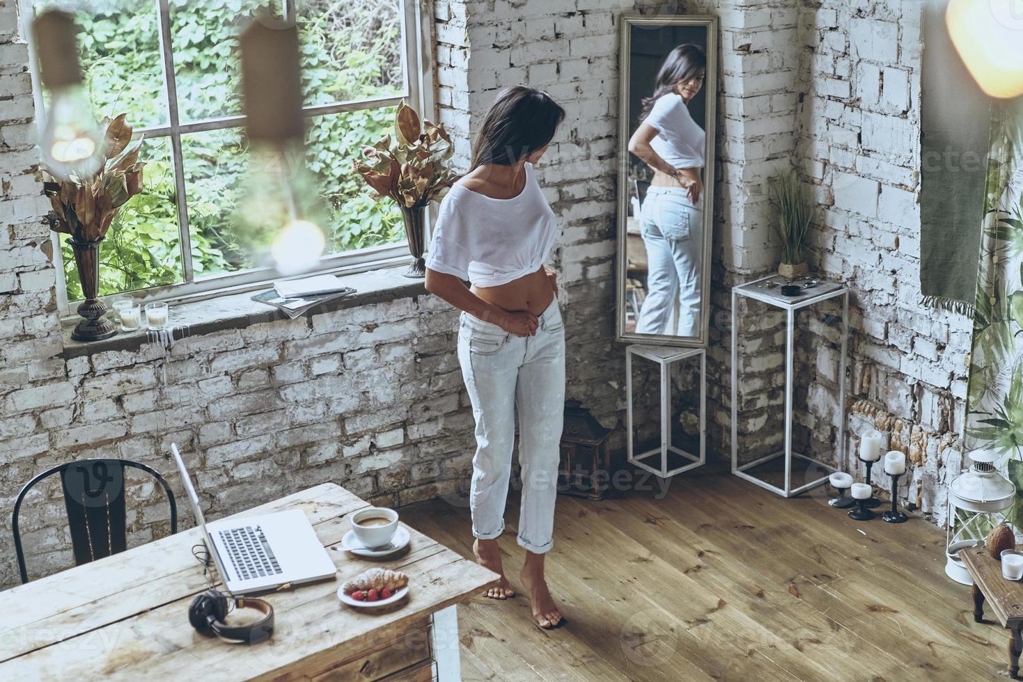 adattamento in magro jeans. pieno lunghezza di attraente giovane donna guardare nel il specchio mentre mettendo su sua jeans foto