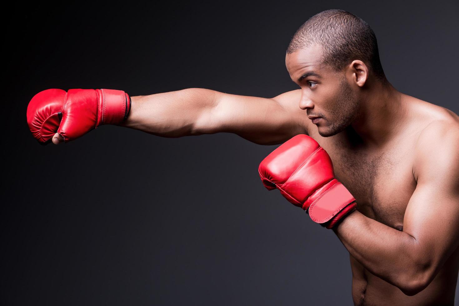 uomo boxe. lato Visualizza di giovane A petto nudo africano uomo nel boxe guanti esercizio mentre in piedi contro grigio sfondo foto