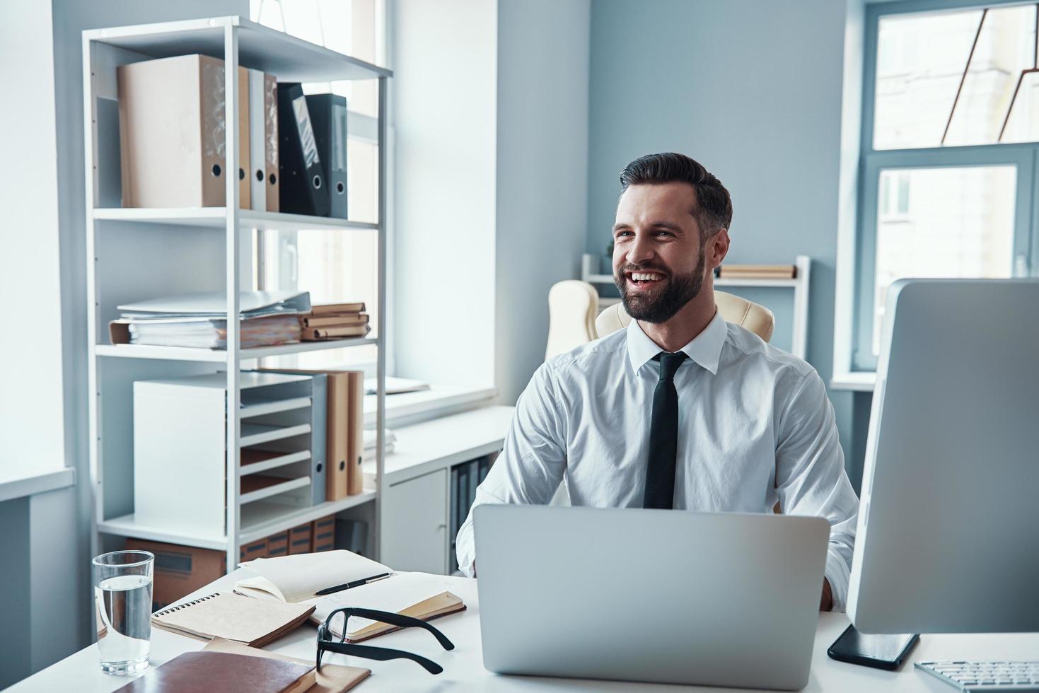 contento giovane uomo nel camicia e cravatta Lavorando utilizzando computer e sorridente mentre seduta nel il ufficio foto