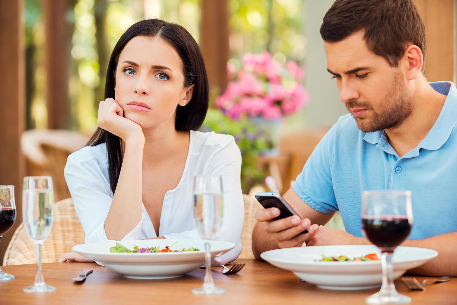 Questo è il peggio Data mai. depresso giovane donna Tenere mano su mento e guardare a telecamera mentre sua fidanzato parlando su il mobile Telefono a all'aperto ristorante foto