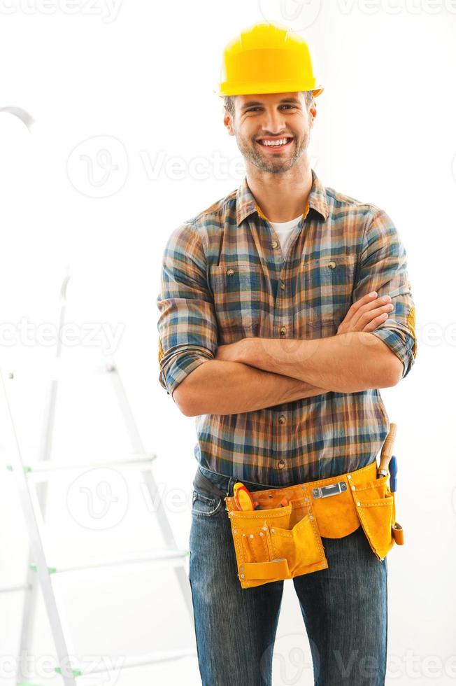 uomo per qualunque lavoro. gioioso giovane costruttore conservazione braccia attraversato e sorridente mentre in piedi in casa foto