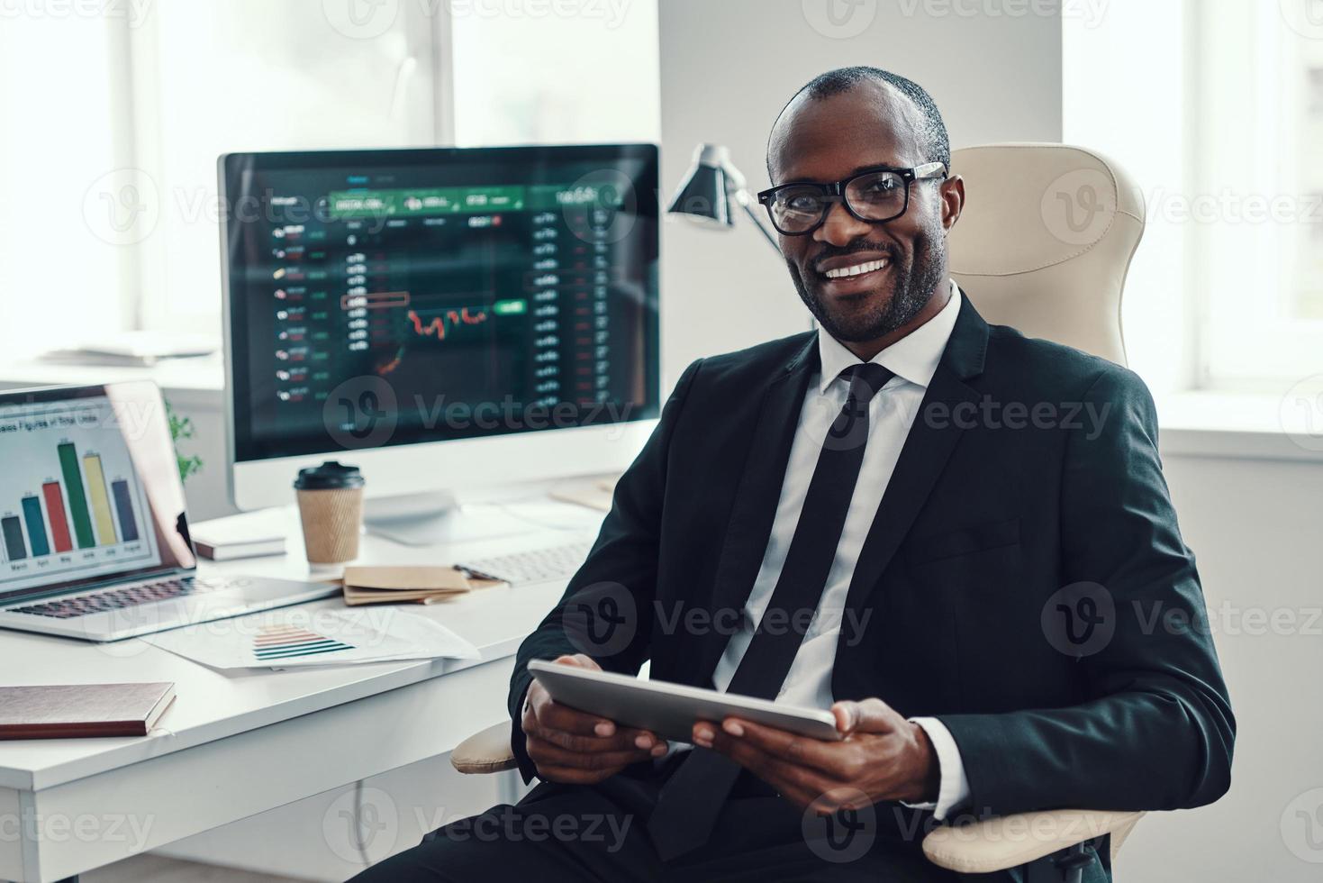 concentrato giovane africano uomo nel vestito formale utilizzando moderno tecnologie e sorridente mentre Lavorando nel il ufficio foto