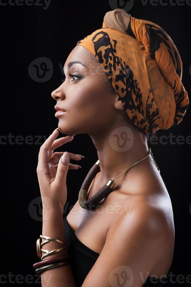 silenzioso bellezza. lato Visualizza di bellissimo africano donna indossare foulard e toccante mento di sua dito mentre in piedi contro nero sfondo foto