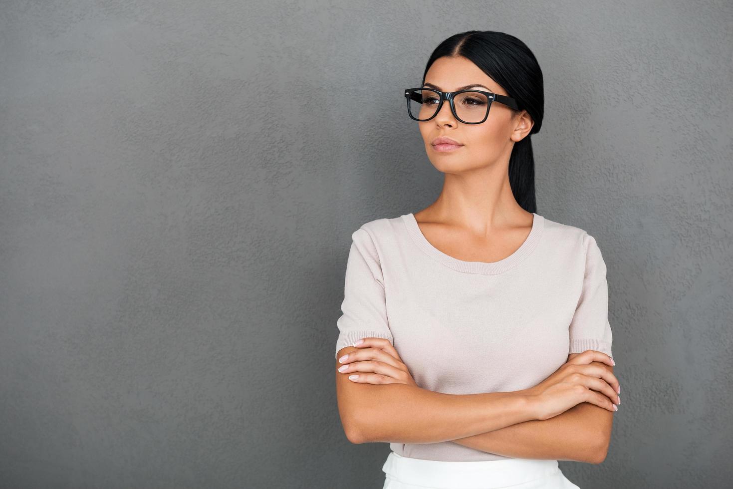 bellissimo e bellissima. fiducioso giovane donna d'affari conservazione braccia attraversato e guardare lontano mentre in piedi contro grigio sfondo foto