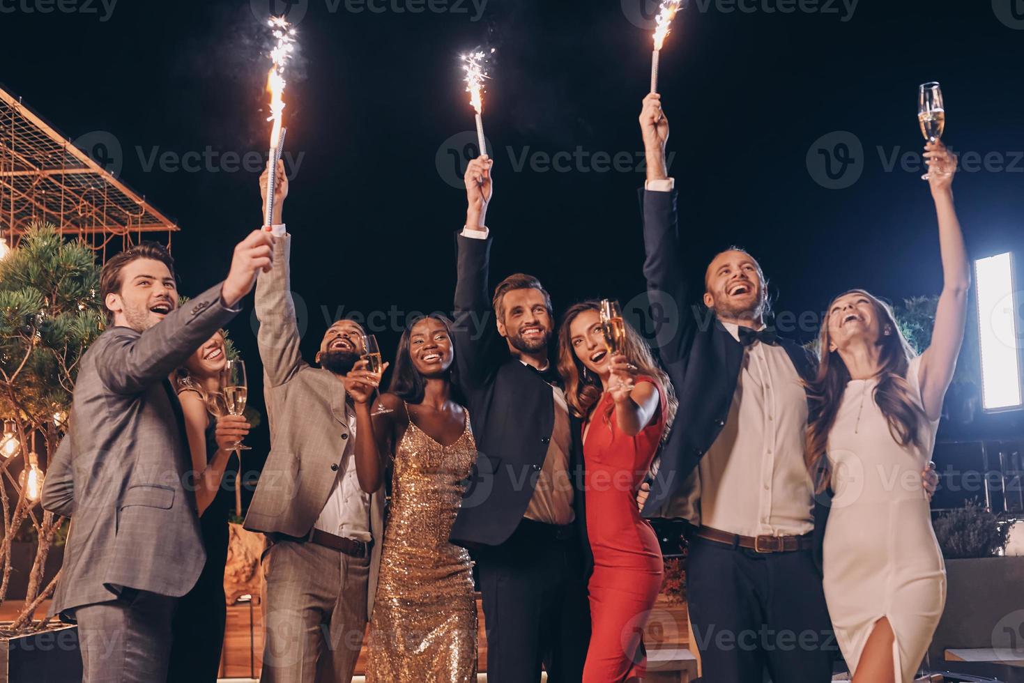 gruppo di bellissimo persone nel vestito formale Tenere sparklers e sorridente mentre la spesa tempo su all'aperto festa foto