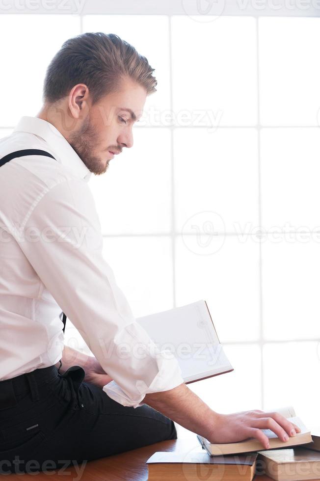 bello topo di biblioteca. posteriore Visualizza di riflessivo giovane uomo nel camicia e giarrettiere seduta su il tavolo e lettura un' libro posa su esso foto