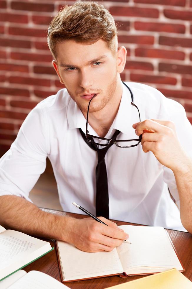 io bisogno alcuni fresco idee. superiore Visualizza di riflessivo giovane uomo nel camicia e cravatta scrittura qualcosa nel Nota tampone e guardare lontano mentre seduta a il suo Lavorando posto foto