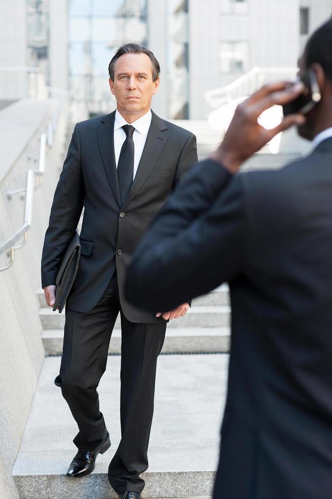 attività commerciale le persone. posteriore Visualizza di africano uomo nel vestito formale parlando su il mobile Telefono mentre un altro uomo d'affari a piedi di le scale su sfondo foto