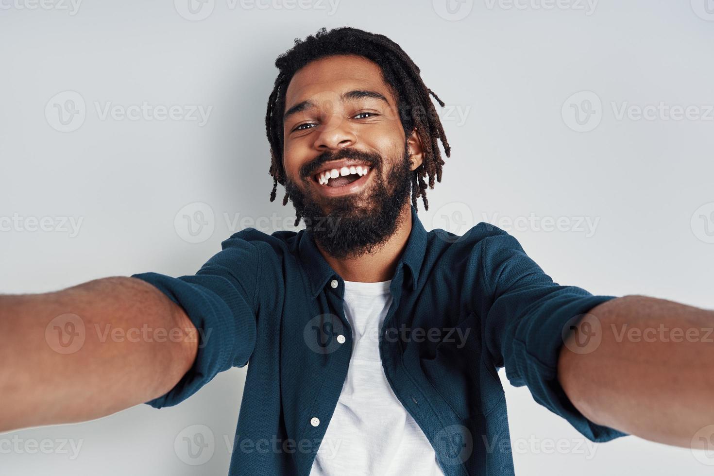 se stesso ritratto di bello giovane africano uomo guardare a telecamera e sorridente mentre in piedi contro grigio sfondo foto