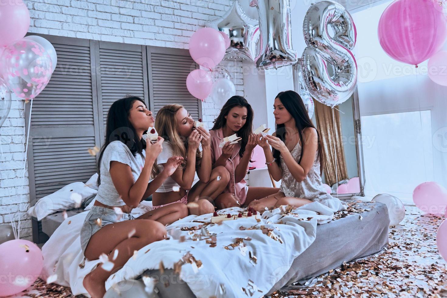 Affamato ragazze. quattro bellissimo giovane donne nel pigiama mangiare torta mentre avendo un' sonno festa nel il Camera da letto con palloncini tutti al di sopra di il posto foto