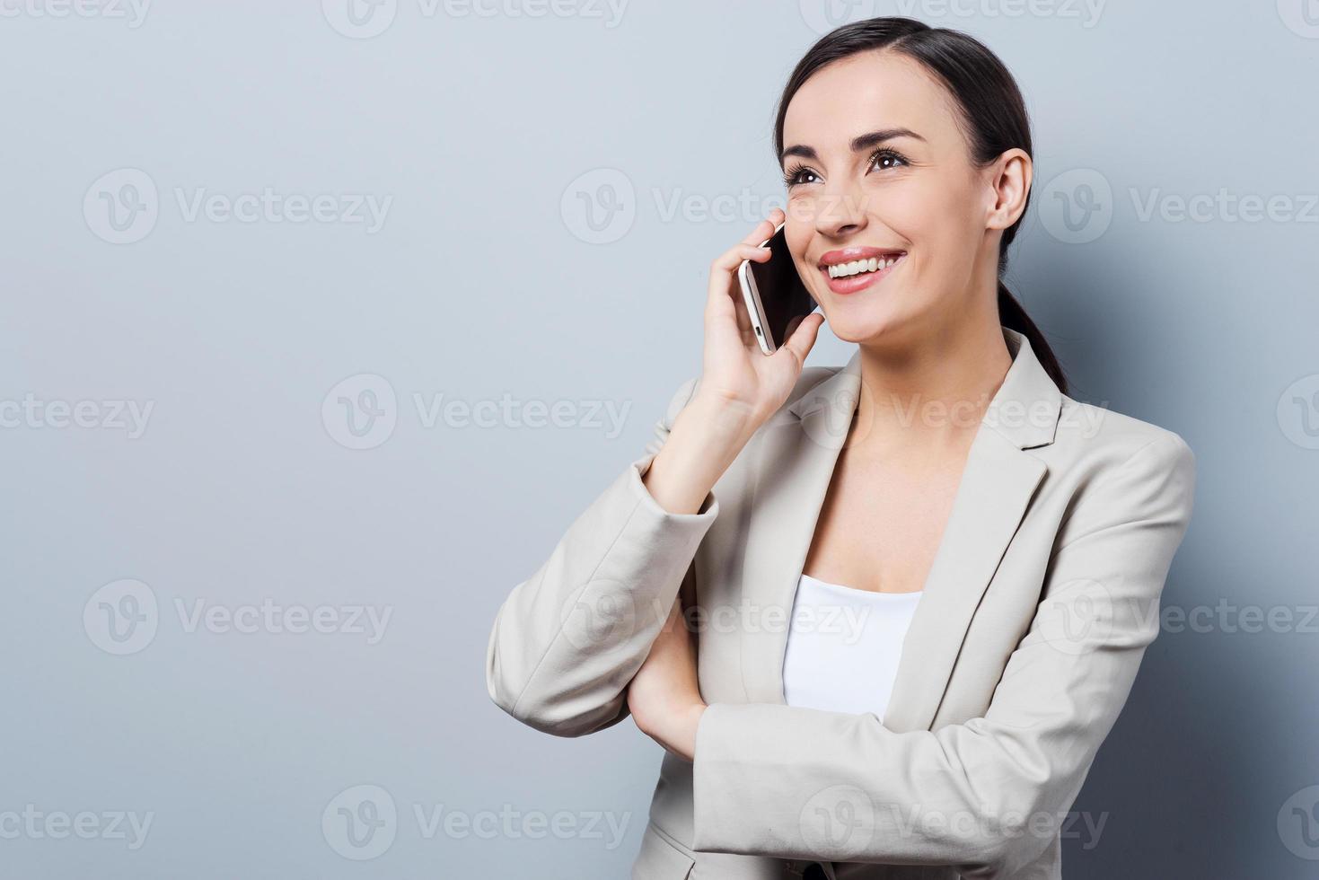 conversazione con piacere. bellissimo giovane donne d'affari parlando su il mobile Telefono e sorridente mentre in piedi contro grigio sfondo foto