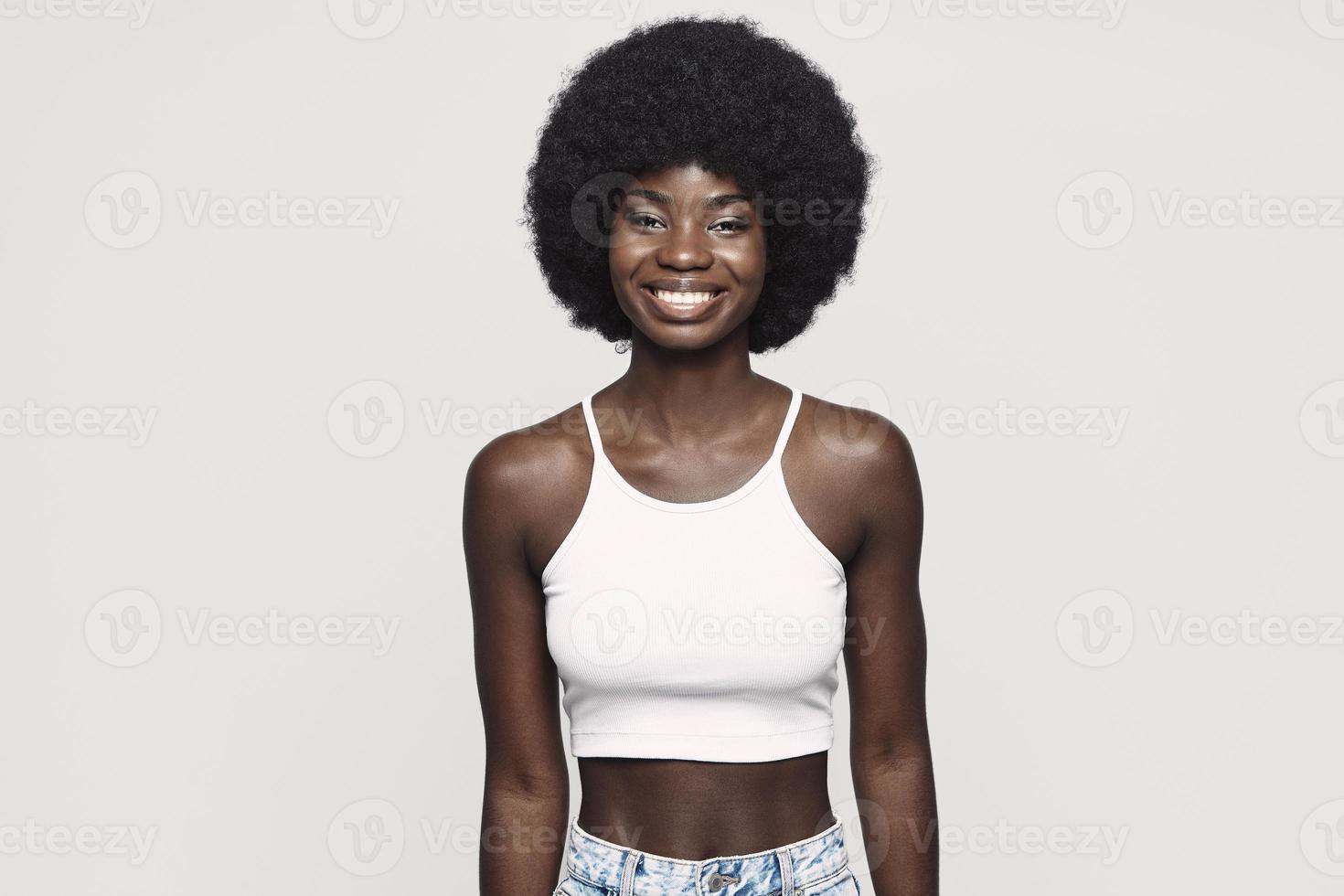 bellissimo giovane africano donna guardare a telecamera e sorridente mentre in piedi contro grigio sfondo foto
