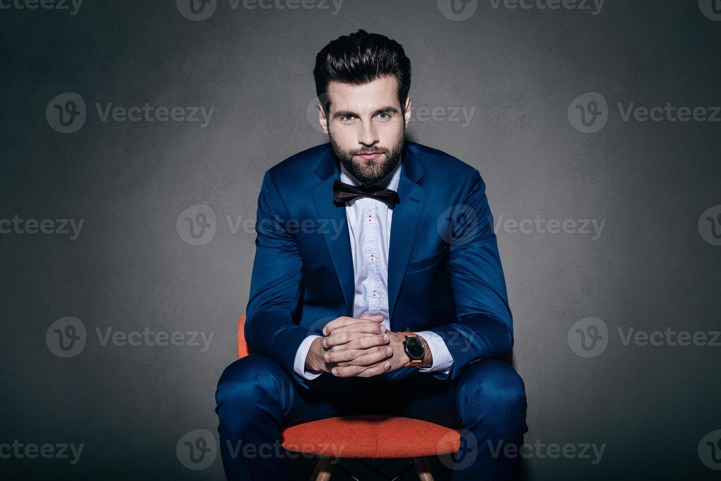 Sig. fiducia. giovane bello uomo nel completo da uomo e arco cravatta guardare a telecamera mentre seduta nel arancia sedia contro grigio sfondo foto