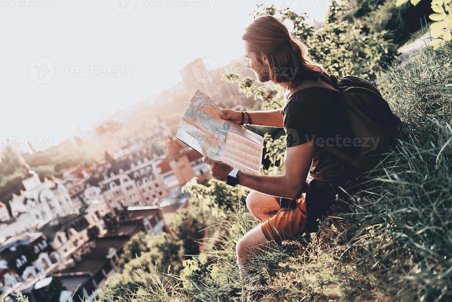 godendo il viaggio. bello giovane uomo nel casuale capi di abbigliamento utilizzando carta geografica mentre seduta su il collina all'aperto foto