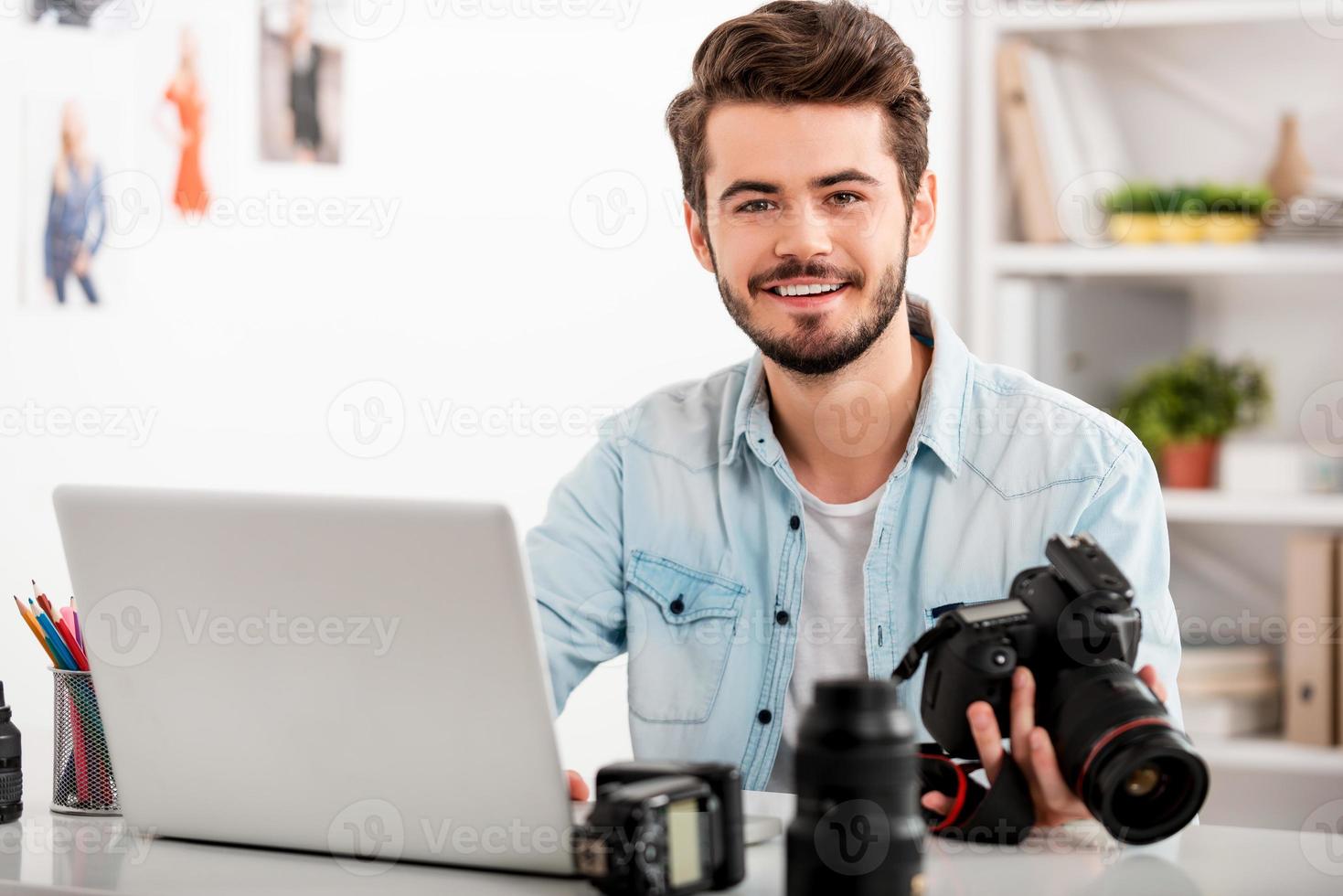 mio lavoro è mio passione. bello giovane uomo Tenere telecamera e sorridente mentre seduta a il suo Lavorando posto foto