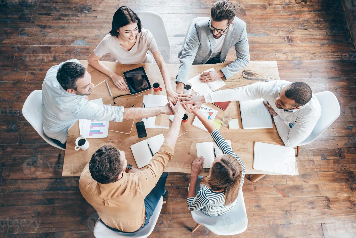 di partenza nuovo Lavorando giorno come un' squadra. superiore Visualizza di gruppo di sei giovane persone Tenere mani insieme e Sorridi mentre seduta a il ufficio scrivania foto