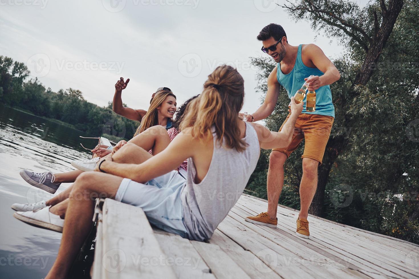 birra volta. gruppo di contento giovane persone nel casuale indossare sorridente e potabile birra mentre seduta su il molo foto