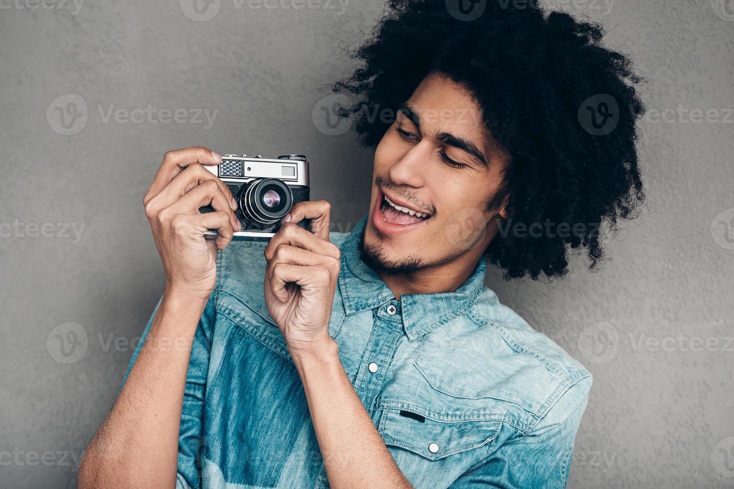 Questo cosa lavori perfettamente bello giovane africano uomo Tenere retrò styled telecamera e guardare a esso con Sorridi mentre in piedi contro grigio sfondo foto