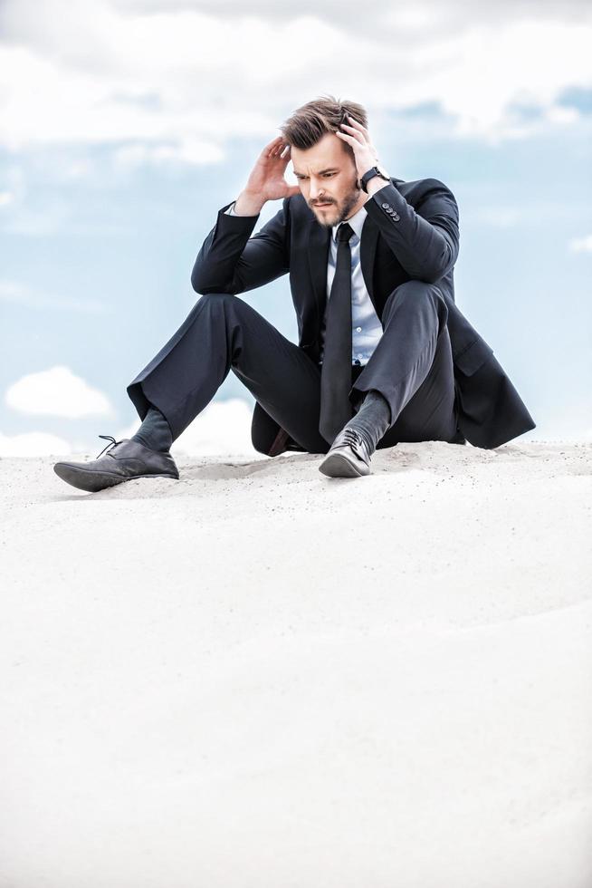 depresso e confuso uomo d'affari. frustrato giovane uomo d'affari Tenere testa nel mani mentre seduta su il superiore di sabbia duna foto