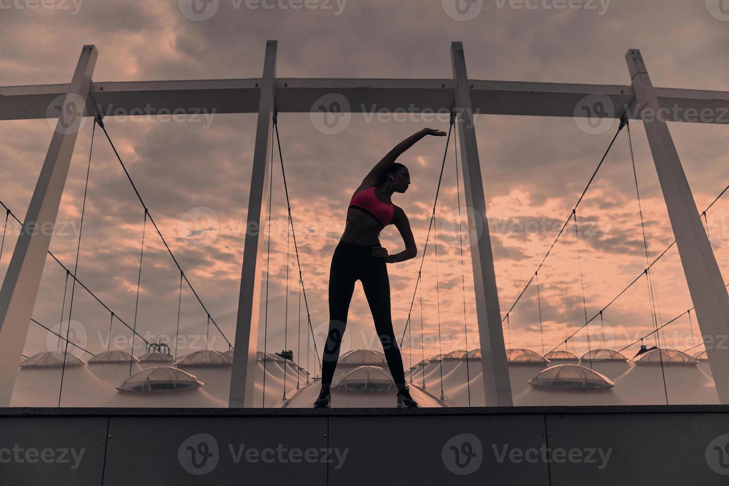 yoga dà energia. pieno lunghezza di moderno giovane donna nel gli sport capi di abbigliamento allungamento mentre riscaldamento su all'aperto foto