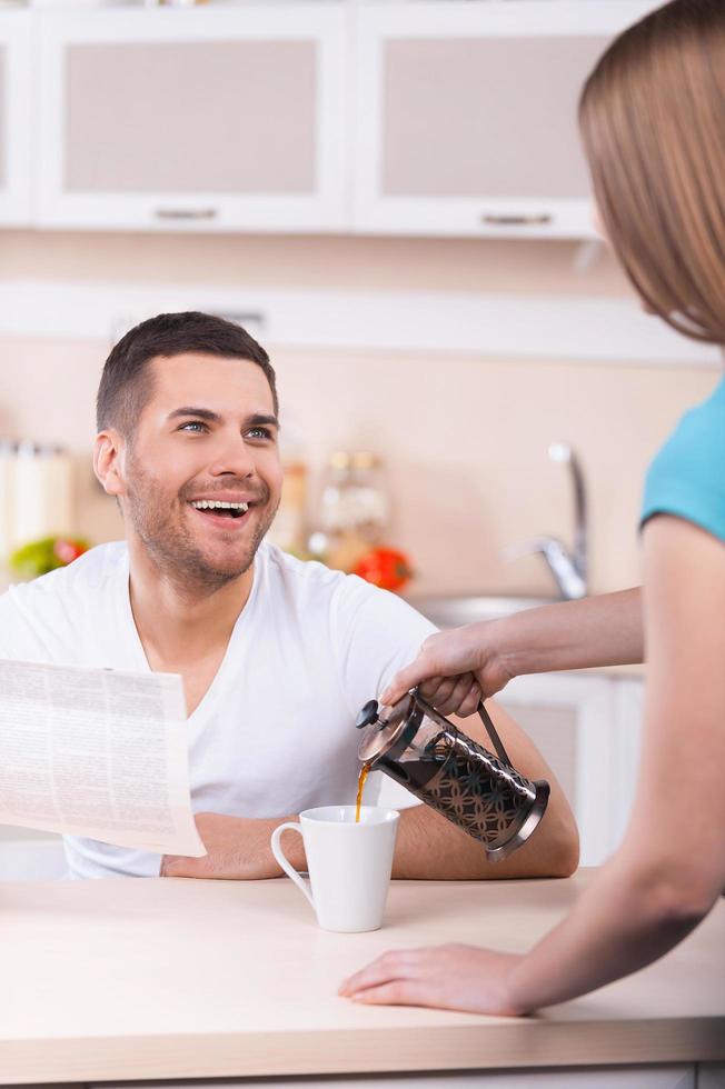 contento Domenica mattina. donna scrosciante caffè per il tazza mentre contento giovane uomo Tenere giornale e sorridente foto