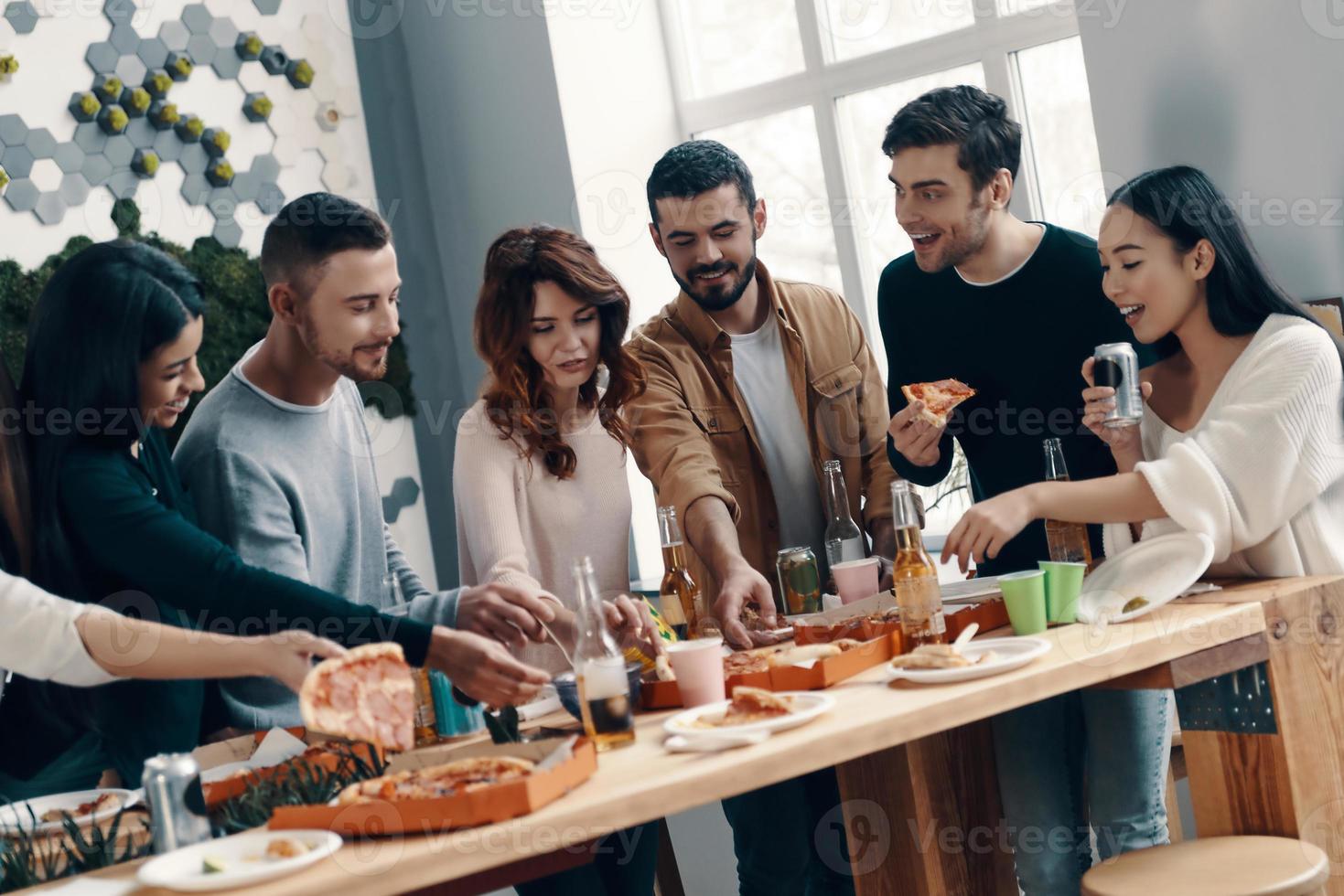 così Affamato gruppo di giovane persone nel casuale indossare mangiare Pizza e sorridente mentre avendo un' cena festa in casa foto