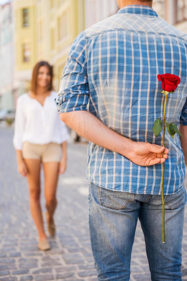 bellissimo fiore per suo. posteriore Visualizza di giovane uomo Tenere rosso rosa dietro a il suo indietro mentre donna a piedi nel il sfondo foto
